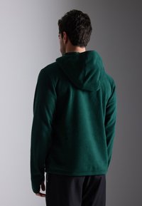 Moleton com capuz em verde escuro com um acabamento texturizado, apresenta um capuz e um corte reto. Costuras visíveis ao longo das bordas.