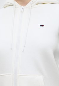 Λευκό φούτερ με φερμουάρ και κορδόνια, με μικρό ορθογώνιο λογότυπο κόκκινου, λευκού και μπλε χρώματος στο αριστερό στήθος.