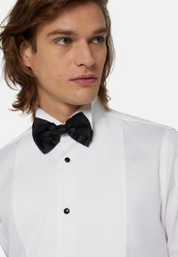 Chemise de smoking blanche avec un devant plissé, accompagnée d'un nœud papillon en satin noir. Comprend deux boutons noirs et un col structuré.