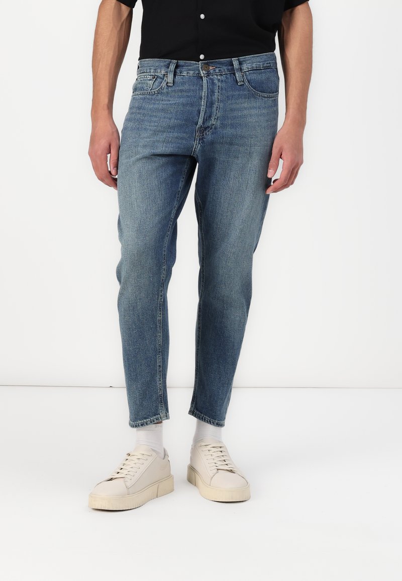 Helle blaue Jeans im geraden Schnitt, mit einem Frontknopfverschluss, fünf Taschen und einem leicht ausgewaschenen Finish. Kombiniert mit beigen Sneakers.