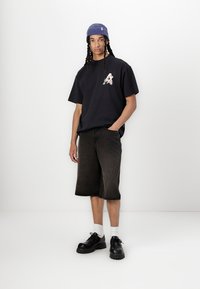 Sort oversized t-shirt med blomster "A" design, parret med mørk denim shorts. Sorte sko og hvide sokker fuldender looket. Blå hat på hovedet.