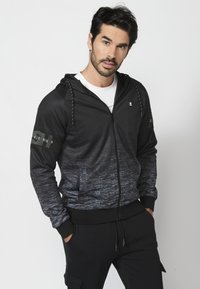 Čierna a šedá gradientná mikina na zips s šnúrkami, bočnými vreckami a logom na rukáve, vyrobená z hladkej, štruktúrovanej tkaniny.