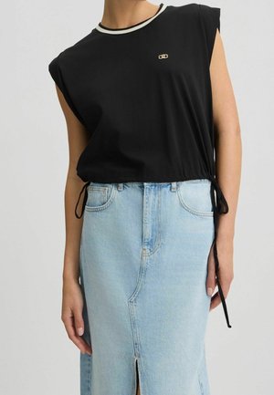 Mujer con top negro sin mangas con ribete blanco y falda vaquera azul con abertura frontal, manos relajadas a los lados.