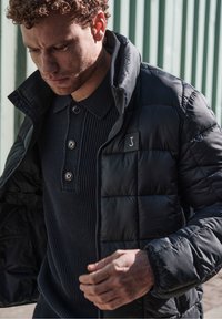 Schwarze Pufferjacke mit gestepptem Muster, getragen über einem strukturierten, marineblauen, gerippten Poloshirt mit drei Knöpfen, das ein geprägtes Logo auf der Jacke aufweist.