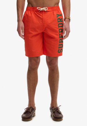 Homme portant un short de bain orange vif avec "SUPERDRY" en lettres vertes sur la jambe gauche, assorti à des chaussures bateau en cuir marron.
