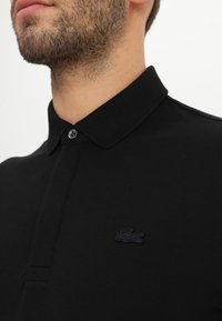 Čierna polo košeľa z bavlnenej látky, s golierom a jedným gombíkom. Výrazné malé čierne logo na ľavej hrudi.