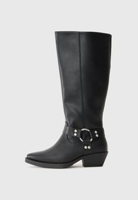 Botas negras de cuero hasta la rodilla con punta afilada, que cuentan con una correa decorativa en el tobillo y detalles de hardware en plata. Tacón bajo y apilado.