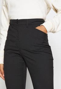 Celana hitam yang pas dengan tekstur halus, pinggang tinggi, saku depan, dan penutup dengan satu kancing. Dipadukan dengan atasan lengan panjang berwarna terang.