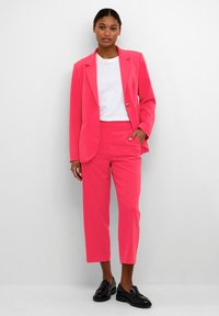 Felle roze pak met een blazer met één knoop en op maat gemaakte broek. De outfit is gecombineerd met een wit t-shirt en zwarte instappers.