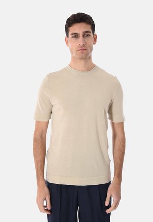 Uomo che indossa un maglione beige a maniche corte di maglia e pantaloni scuri, in piedi contro uno sfondo chiaro uniforme, rivolto in avanti con un'espressione neutra.