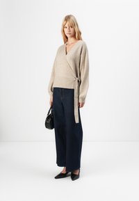 Blond kvinna iklädd en beige omlottkofta, mörka vida jeans, svarta spetsiga skor och håller en liten svart handväska framför en vit bakgrund.