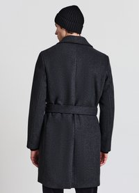 Cappotto in lana grigio scuro con collo strutturato, dettaglio cinturino posteriore e texture sottile. Indossato con un berretto lavorato a maglia nero.