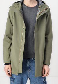Voděodolná bunda olivově zelené barvy s kapucí, předním zipem, šedou vnitřní podšívkou a nastavitelnými šňůrkami, doplněná šedým svetrem pod ní.