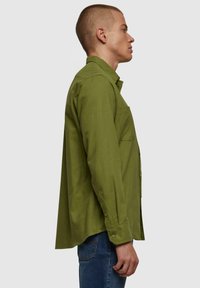 Camisa verde oliva de botones, con un bolsillo en el lado izquierdo, hecha de una tela texturizada, con un corte recto y mangas con puños.