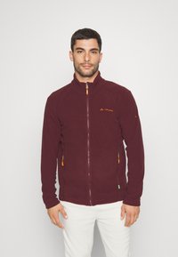 Giacca in fleece bordeaux con colletto alto, cerniera frontale e tasche laterali con tirazip arancioni. Logo sul lato sinistro del petto.
