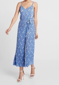 Modrý květinový overal s tenkými ramínky, výstřihem do V, nastavitelným páskem a širokými nohavicemi. Vyroben z lehké tkaniny, zdobený jemnými květinovými vzory.