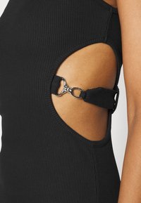 Černý ribovaný oděv s otevřenými bočními stranami a nastavovacími popruhy upevněnými stříbrnou sponou. Hladká textura s dobře definovaným švem.