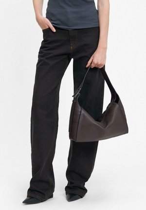 Borsa a spalla in pelle marrone con un design morbido e tracolla regolabile, tenuta in mano. Modella che indossa pantaloni larghi neri e un top aderente.