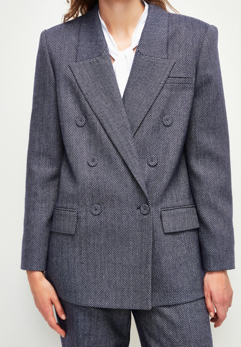 Blazer de doble botonadura en patrón espiga gris, con solapas de muesca, bolsillos angulados y hombros estructurados. La tela tiene una textura suave.