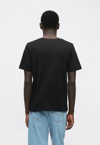 Mann med korte krøller iført svart t-skjorte og lyseblå jeans, sett bakfra mot en ensfarget hvit bakgrunn.