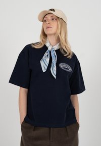 Femme blonde portant une casquette beige, un t-shirt oversize bleu marine avec un patch, une écharpe bleue à motifs nouée autour du cou, et un pantalon marron, les mains dans les poches.
