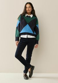 Argyle-mønstret sweater i grøn, blå og sort, parret med hvid skjorte med krave og mørkeblå bukser, afsluttet med blanke sko.