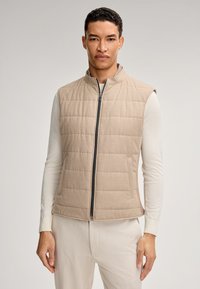 Beige gesteppte Weste mit hohem Kragen und Reißverschluss; weiche Textur und Seitentaschen. Über einem hellen Langarmhemd getragen.