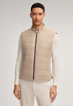 Gilet trapuntato beige con alto colletto e zip; texture morbida e tasche laterali. Indossato sopra una camicia a maniche lunghe di colore chiaro.