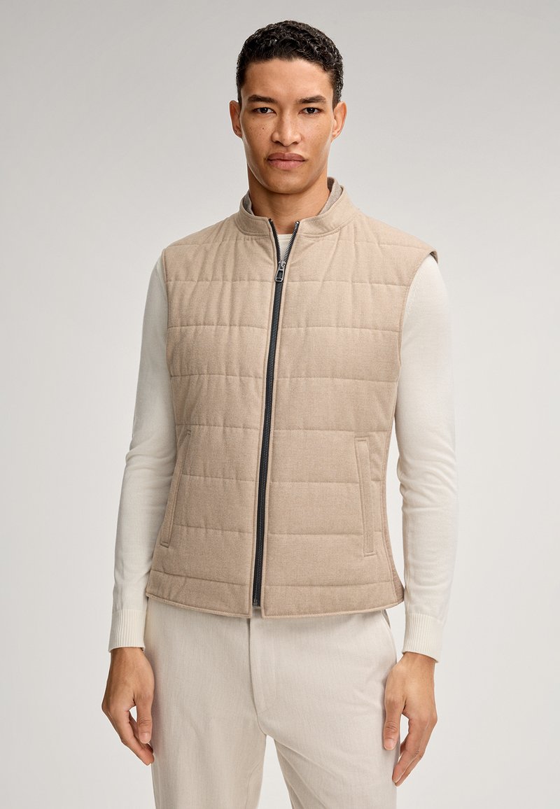 Beige gesteppte Weste mit hohem Kragen und Reißverschluss; weiche Textur und Seitentaschen. Über einem hellen Langarmhemd getragen.