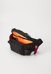 Svart midjeväska med orange innside, frontlomme med glidelås, justerbar stropp, og rød tekstilmerke med hvit logo festet til glidelåsen.