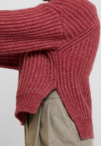 Pull en tricot de couleur rose profond, avec une texture côtelée, des fentes sur les côtés et une coupe décontractée. Le matériau semble doux et chaud.