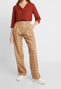 Rust-colored collared blouse paired with tan plaid trousers featuring a high waist and side pockets, completed with white sneakers.