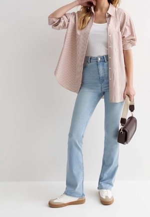 Femme portant un jean taille haute bleu clair, un débardeur blanc, une chemise oversize à rayures roses, des baskets blanches, tenant un petit sac à main marron.