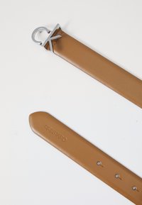 Ceinture en cuir tan ornée d'une boucle circulaire en argent avec le logo "CK", texture lisse, et cinq trous d'ajustement le long de la sangle.