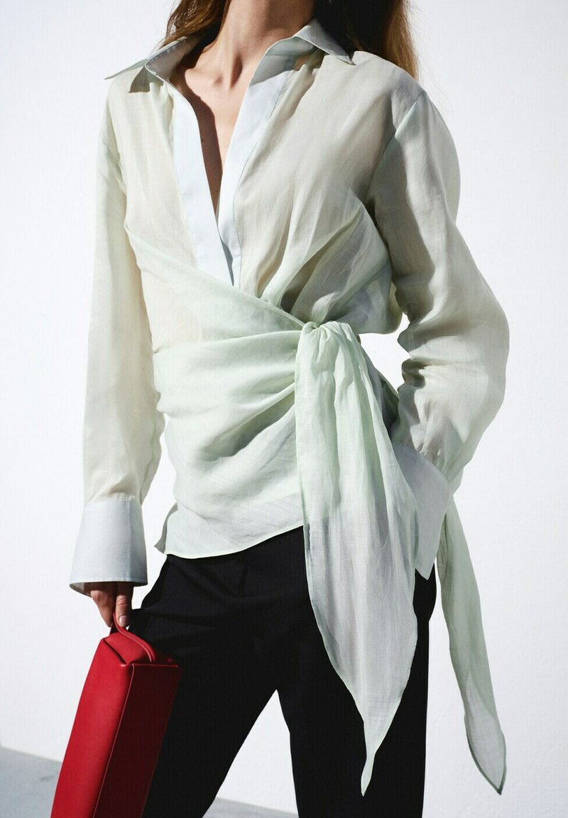 Femme portant un chemisier léger transparent par-dessus une chemise blanche, avec un pantalon noir, tenant un sac à main rouge rectangulaire sur fond blanc.