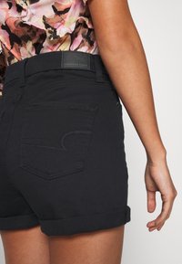 Femme portant un short en jean noir à ourlet roulé et un haut rose à motif floral, vue de la taille jusqu'en bas avec le bras détendu le long du corps.