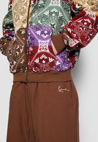 Multicolored zip-up jacket with intricate floral patterns, brown cuffs; paired with brown pants featuring a white embroidered logo.