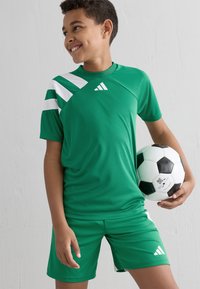 Niño sonriendo y sosteniendo un balón de fútbol en blanco y negro, vestido con un conjunto deportivo verde de Adidas frente a un fondo blanco liso.