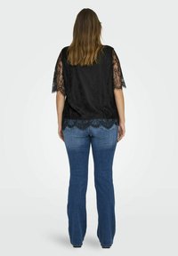 Frau, die mit dem Rücken zur Kamera steht und ein schwarzes Spitzenoberteil mit kurzen Ärmeln sowie helle, ausgebogene Jeans trägt, vor einem hellen Hintergrund.
