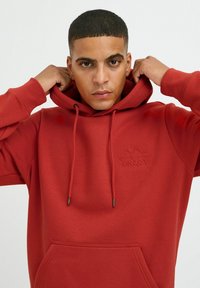 Red hoodie made of textured fabric, featuring a front pocket, drawstring hood, and embossed "ENJOY" logo detail.
