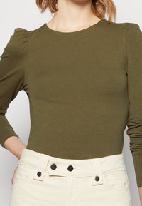 Frau trägt ein olivgrünes Langarmoberteil mit gepufften Schultern und hochgeschnittene, cremefarbene Hose mit sichtbaren schwarzen Knöpfen.