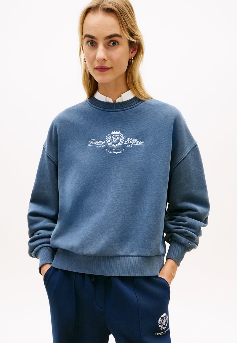 Γυναίκα με μπλε φούτερ Tommy Hilfiger και αντίστοιχο παντελόνι φόρμας, στέκεται με τα χέρια στις τσέπες μπροστά σε απλό φόντο.