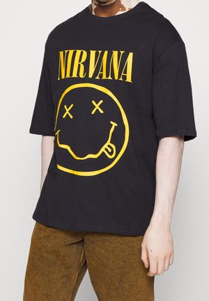 Osoba nosząca czarną koszulkę Nirvana z żółtym logo uśmiechniętej buzi i krzyżykami zamiast oczu, połączoną z brązowymi spodniami.