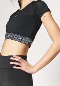 Idrottare iklädd svart Nike Pro cropped top med meshärmar och högmidjade svarta leggings, stående med handen på höften.