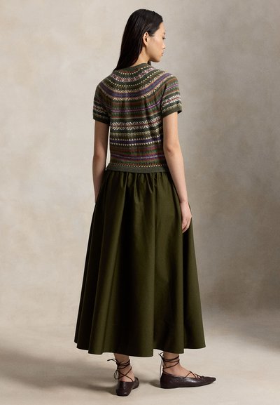Femme portant un pull à manches courtes à motifs, une longue jupe vert foncé et des chaussures plates à lacets foncées, debout de dos à la caméra.