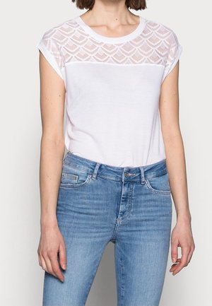 Biała koszulka z krótkim rękawem z teksturowaną koronkową wstawką na ramionach, zestawiona z jasnoniebieskimi dżinsami. Materiał wydaje się miękki i gładki.