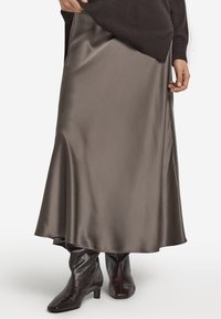 Satynowa długa spódnica w stonowanym odcieniu taupe, charakteryzująca się gładką fakturą i płynnym kształtem, zestawiona z ciemnobrązowymi botkami na obcasie.