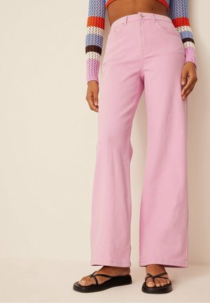 Jeans évasés en denim rose, avec une taille haute, une fermeture à bouton classique et des détails de couture subtils. Associés à des tongs noires.