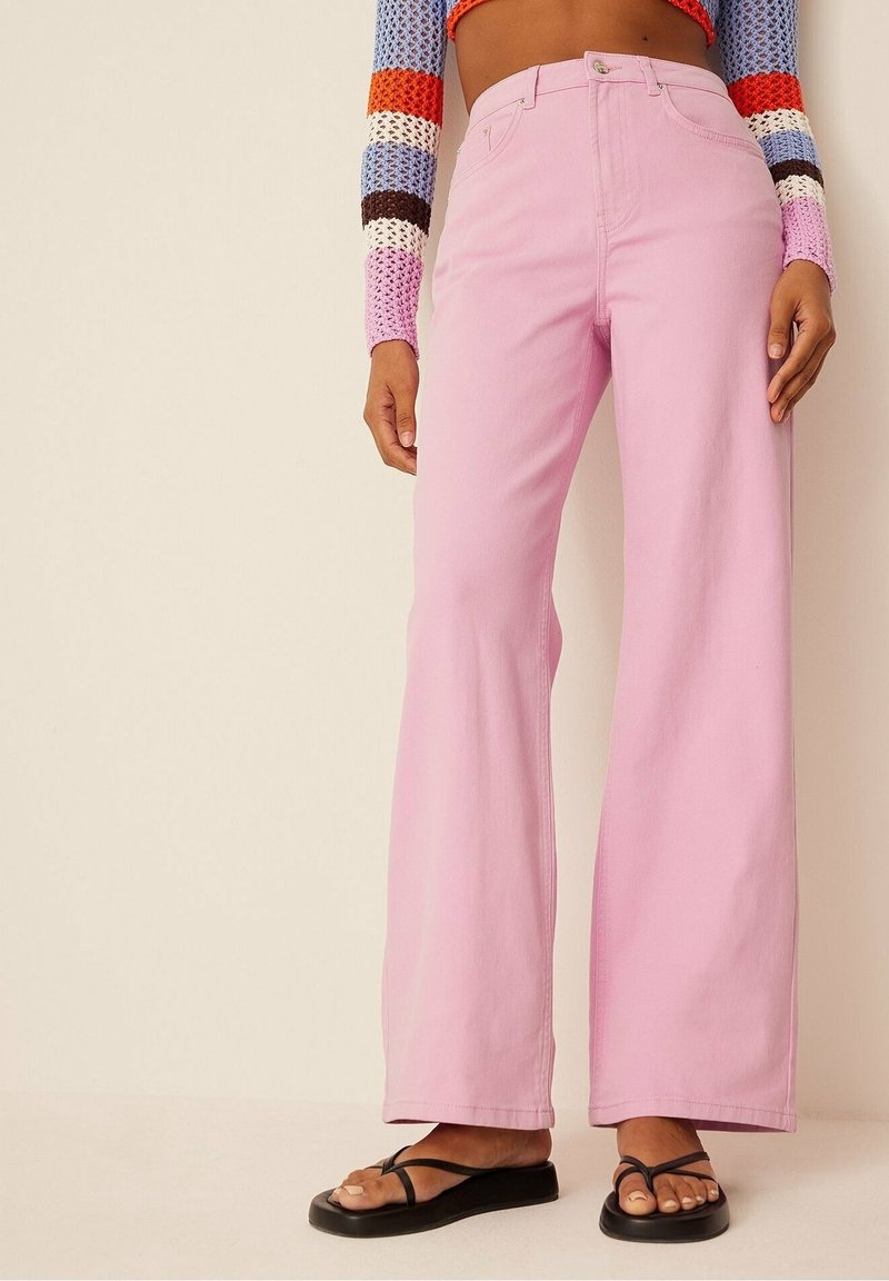 Vidbenade jeans i rosa denim med hög midja, klassisk knappstängning och subtila sömnadsdetaljer. Kombinerat med svarta flip-flops.