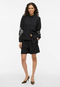 Camicia nera con bottoni e maniche lunghe con ricami floreali sulle maniche, abbinata a pantaloncini neri e tacchi verdi a punta.